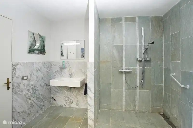 Baño de mármol con ducha, inodoro y lavabo. Hay barras de apoyo fijadas a la pared para discapacitados y hay disponible una silla de ruedas para la ducha.