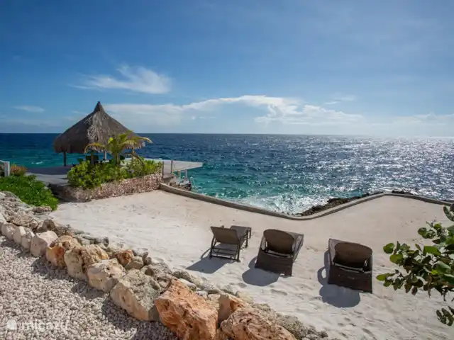 Villa frente al mar en el resort Boca Gentil en Curazao, Banda Arriba (este), Jan Thiel - villa La villa con vistas desde la gran terraza de la piscina. Ves la palapa con la terraza flotante en el mar. La playa ajardinada lo completa. Aquí puedes tomar el sol y estar cerca del agua.