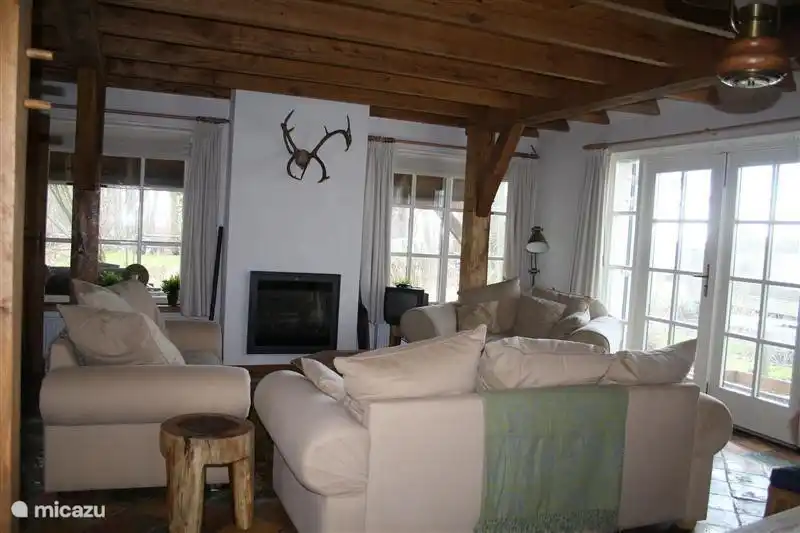 De gezellige woonkamer met zit rondom de (electrische) openhaard. Er is een tv en voldoende licht om een boek te lezen. Mocht u liever buiten willen zitten, via de openslaande deuren komt u op het terras met houten stoelen en tafel uitkijkend over de tuin, water en de Weerribben-Wieden