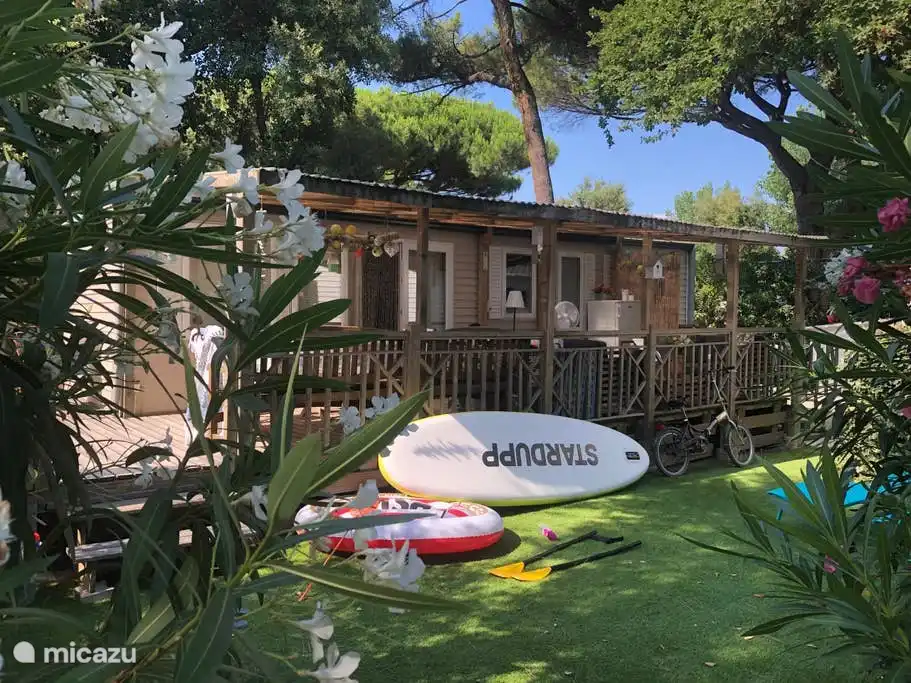 Nuestra casa móvil, en parte a la sombra bajo los árboles, en parte con el sol en la terraza y el césped frente a ella. Plaza de aparcamiento delante de la casa móvil.