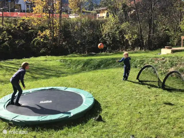 Berghaus Christel en Austria, Carintia, Arnoldstein - casa vacacional Vista desde la terraza en el jardín. Disfruta del trampolín, juega al fútbol, al bádminton o relájate bajo el sol. Hay una cabaña en el bosque en la parte trasera del jardín. A esto se puede llegar a través de una pasarela sobre el arroyo. ¡Puedes construir bonitas represas en el arroyo!