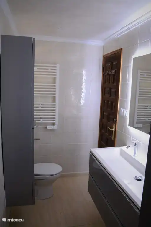 Badezimmer von der Dusche aus gesehen mit Doppelwaschbecken, beleuchtetem Spiegel, Schrank, WC und (Handtuch-)Heizung.