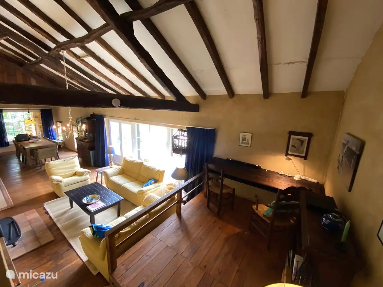 Das Wohnzimmer mit Sitzecke, Esstisch von der anderen Seite des Raumes gesehen. Der Raum ist ca. 16 Meter lang. Rechts die große Terrasse auf der Talseite. Das Foto wurde von einer Anhöhe aus aufgenommen, es gibt ein Bücherregal und einen Schreibtisch, der von 2 Personen genutzt werden kann.