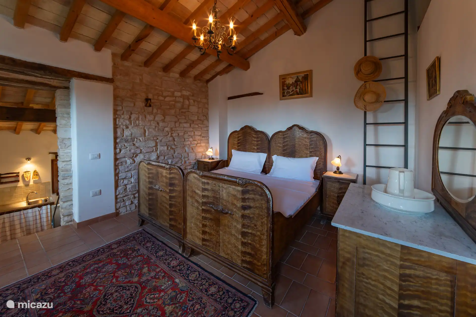 sfeervolle slaapkamer met een uniek, handgemaakte houten tweepersoonsbed. Prachtige houten balkenplafond, kroonluchter en natuurstenen muur. Warme romantische sfeer in het hart van Le Marche. 