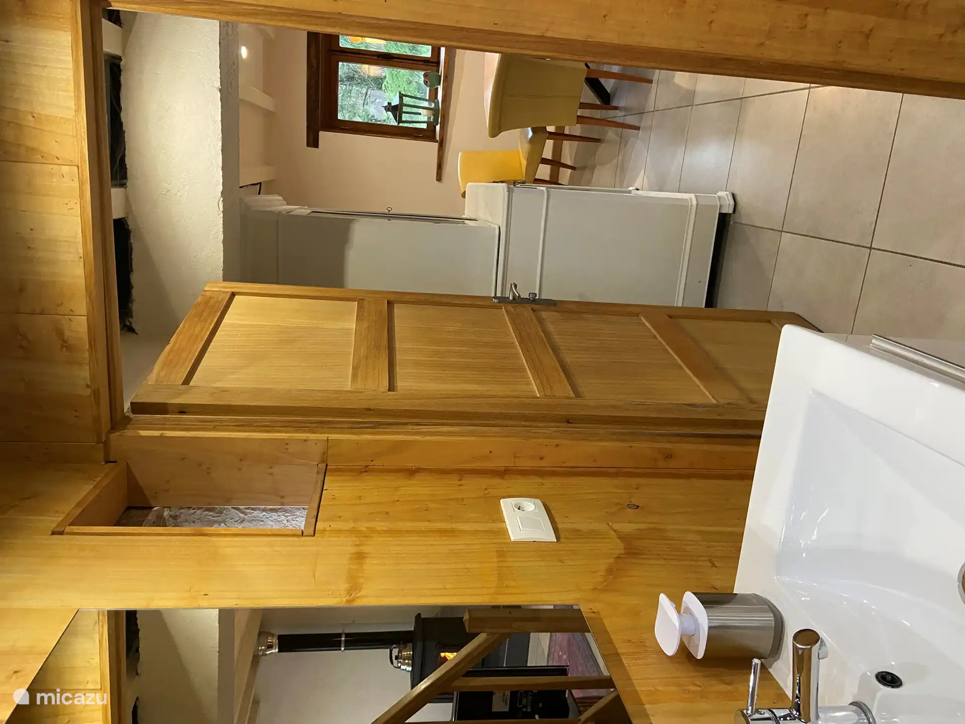 Der Holzofen spiegelt sich im Spiegel des Badezimmers. Im Zimmer sieht man den Esstisch und das Fenster mit Blick auf den Garten.