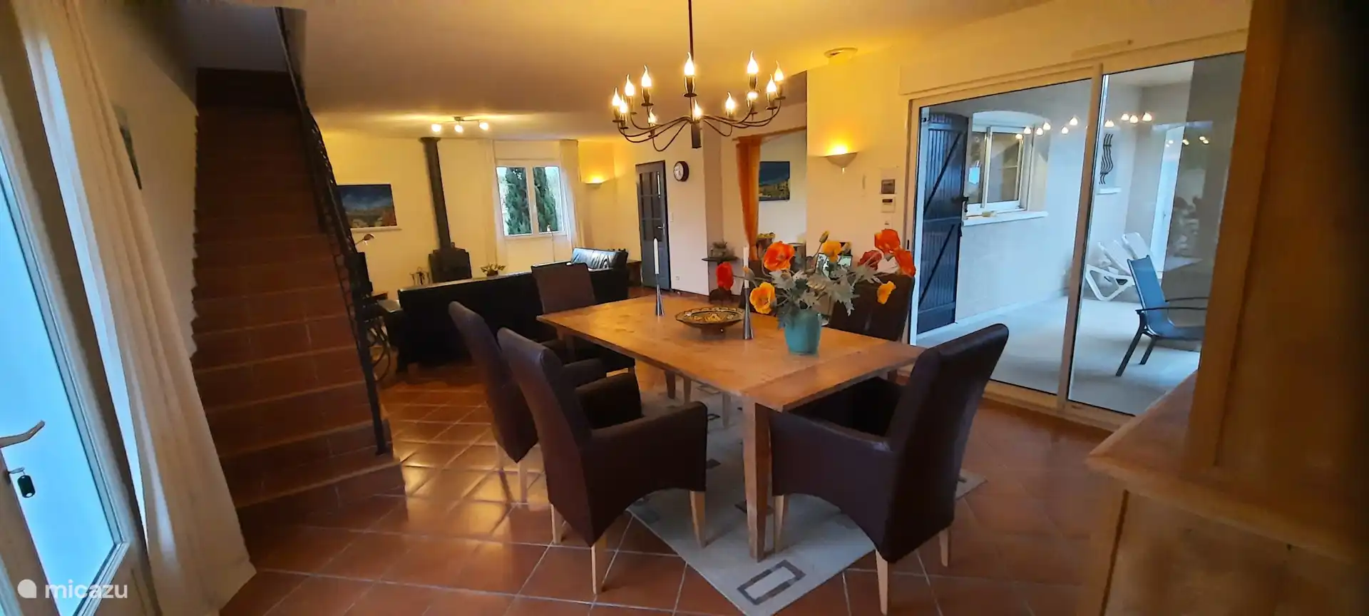 Salle à manger avec grande table avec 6 chaises. Portes coulissantes donnant sur la terrasse et un escalier en pierre menant à la tour chambre avec bibliothèque et canapé-lit. Devant, l’entrée de la cuisine ouverte et la porte du hall.