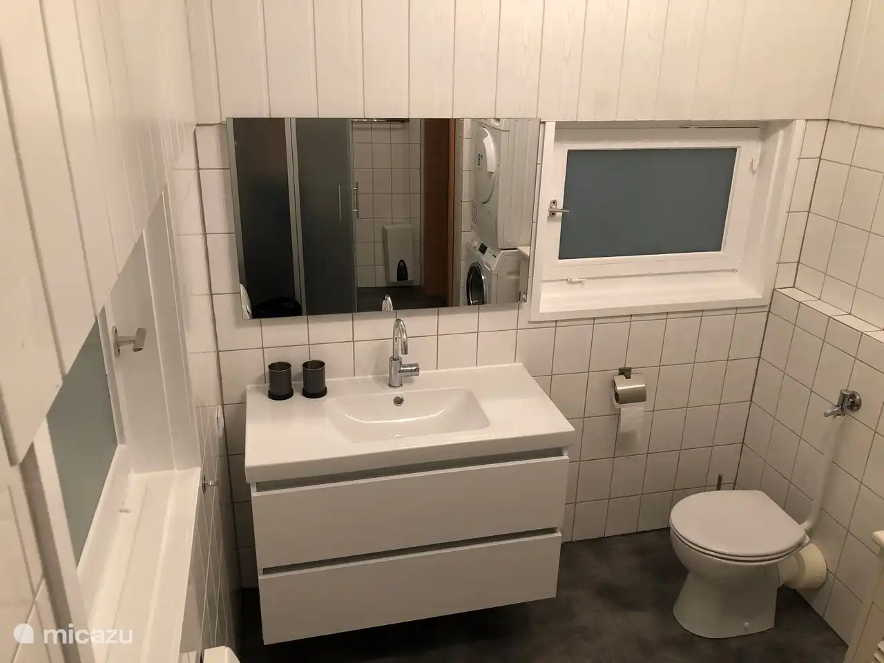 La salle de bain a un lavabo avec miroir, WC, douche spacieuse et machine à laver et sèche-linge.