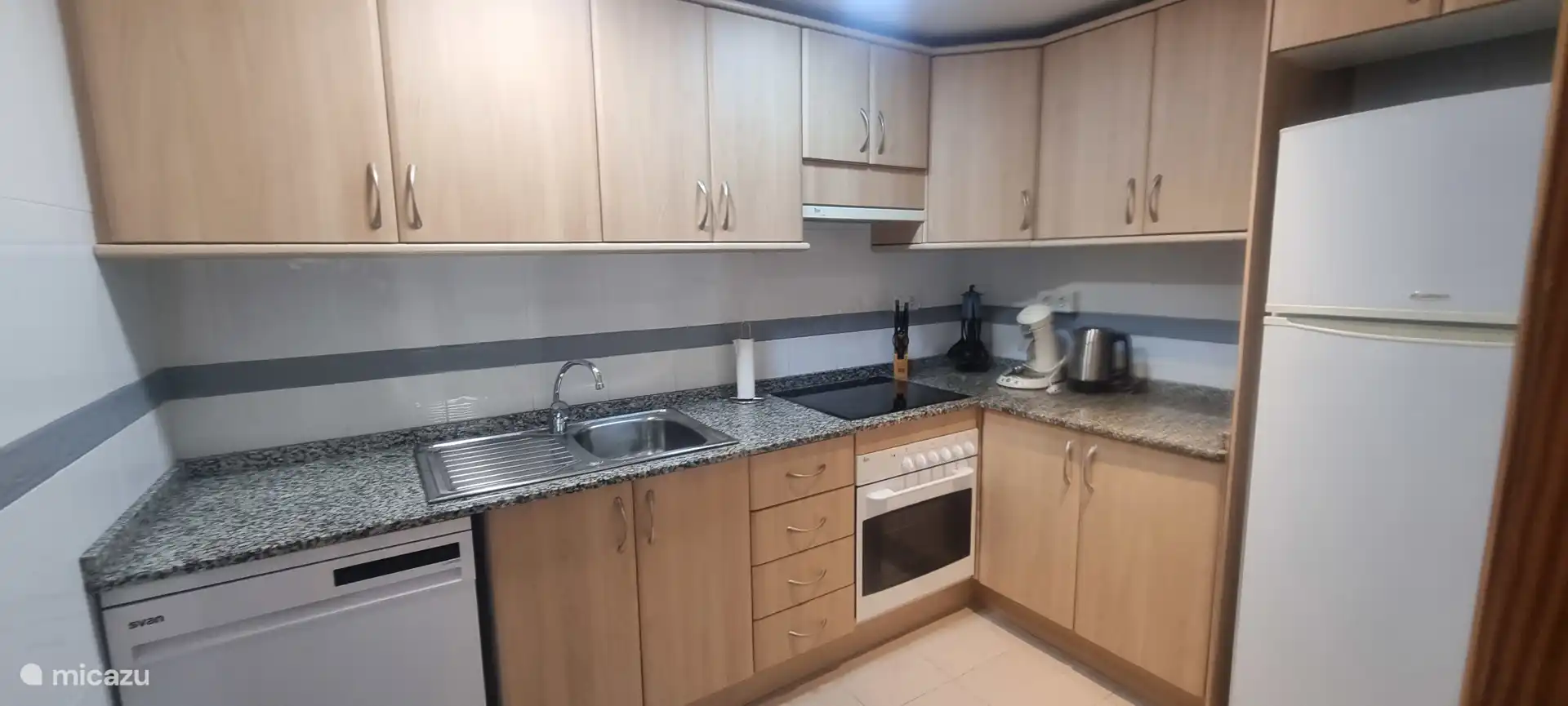 Die Küche mit Induktionsplatte, großem Kühlschrank mit separatem Gefrierschrank, neuer Geschirrspüler und verschiedenen Küchengeräten. Angrenzend befindet sich ein separater Hauswirtschaftsraum mit Waschmaschine und Waschbecken.