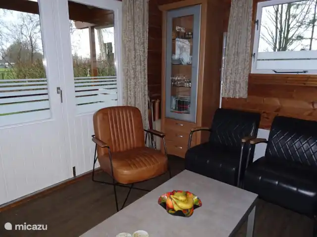 Chalet finlandais, dans un parc avec piscine | Pays-Bas, Overijssel, Gramsbergen - maison de vacances Salon confortable avec portes-fenêtres donnant sur la véranda. Il y a aussi une vitrine, où l’on peut trouver des livres et de la vaisselle. Les tiroirs contiennent des jeux et des livres.