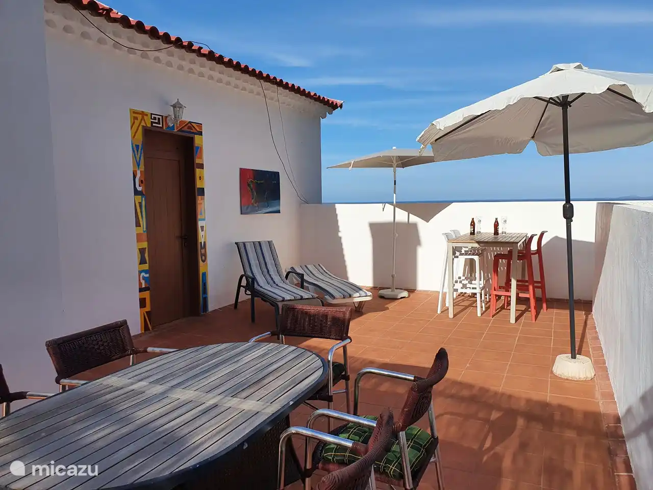 La gran terraza con 2 tumbonas con cojines, zona alta de asientos con taburetes de barra. puerta al dormitorio principal con vistas al mar y televisión