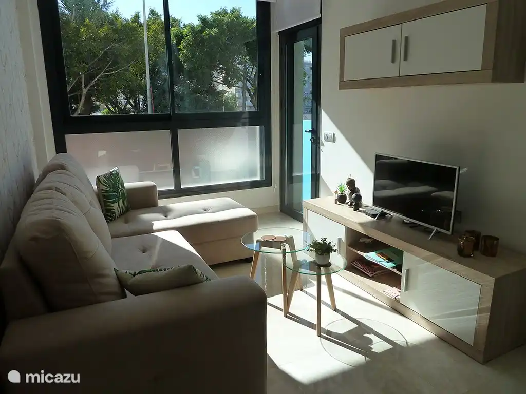 Schön ausgestattetes Wohnzimmer mit Blick auf einen Garten. TV Flandern und WLAN vorhanden. Die Wohnung ist außerdem mit Klimaanlage und elektrischen Rollläden ausgestattet.