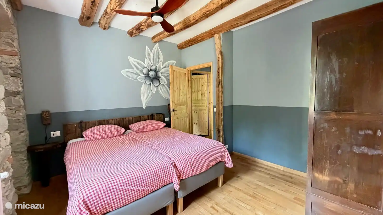 Slaap heerlijk in de sfeervolle slaapkamer van Appartement Stella in Valsesia, Piemonte. Met comfortabele boxspringbedden, een plafondventilator, authentieke houten balken en warme, rustieke details.
