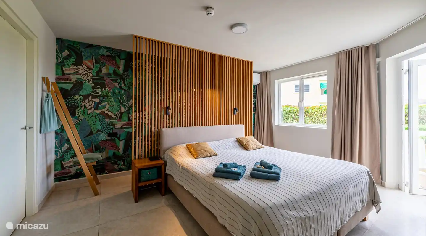 La chambre avec un lit avec sommier à ressorts king-size et un poste de travail derrière le mur à lattes. La chambre est adjacente à la salle de bain et au walk-in. Les serviettes de bain et de plage sont prêtes ici.