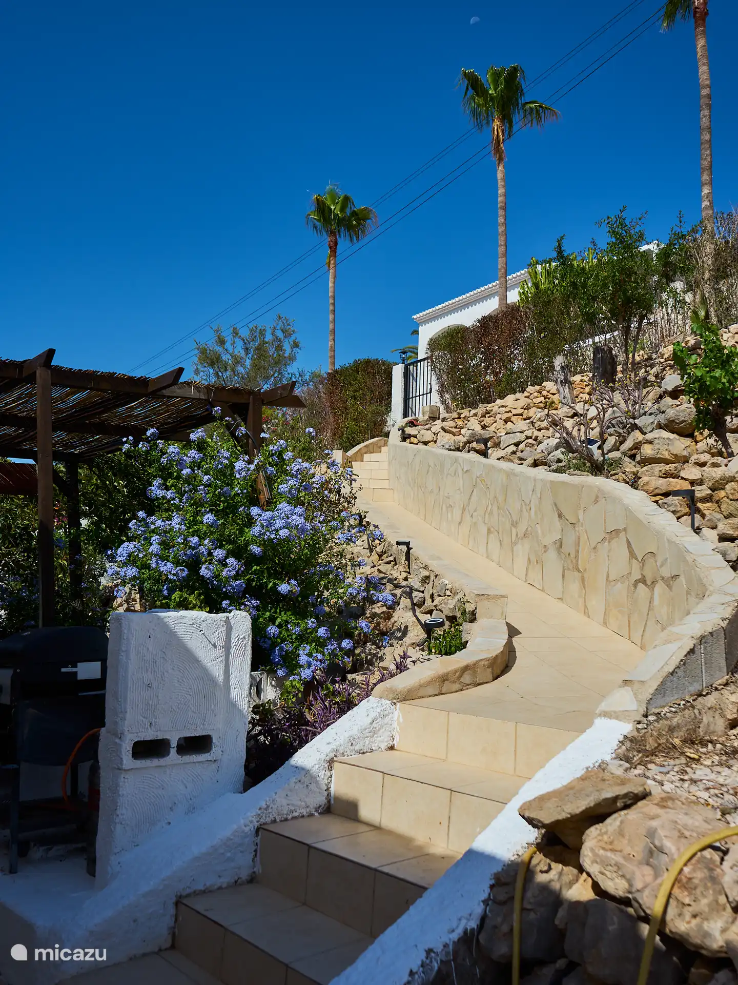 Blick von der Haustür der Casa Yavari
Von der Haustür aus blickst du auf das höhere Tor. Auf der linken Seite sehen Sie die Pergola im Ibiza-Stil mit zwei schwarzen Ibiza-Lampen, die von üppigen Blumen umgeben sind. Ebenfalls zu sehen ist der Grill, der perfekt in diesen attraktiven Außenbereich integriert ist.