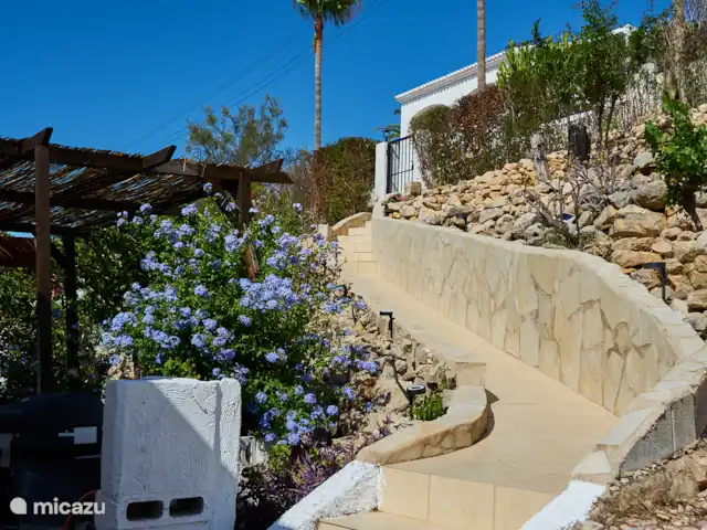 Casa Yavari en España, Costa Blanca, Pedreguer  - villa Vista desde la puerta principal de Casa Yavari
Desde la puerta principal se mira hacia la puerta superior. A la izquierda, se puede ver la pérgola de estilo ibicenco con dos lámparas negras ibicencas, rodeadas de exuberantes flores. También es visible la barbacoa, perfectamente integrada en este atractivo espacio exterior.