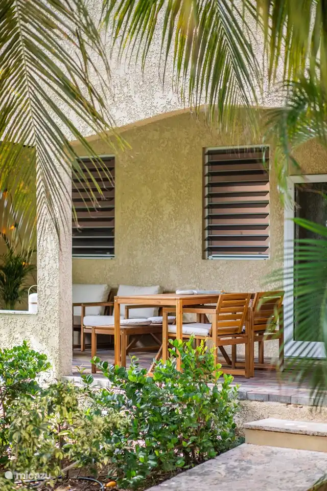 Porche spacieux au rez-de-chaussée avec accès direct à la piscine, abrité par des plantes tropicales et des arbres. Comprend un coin salon confortable, une table à manger et un ventilateur de plafond pour plus de confort
