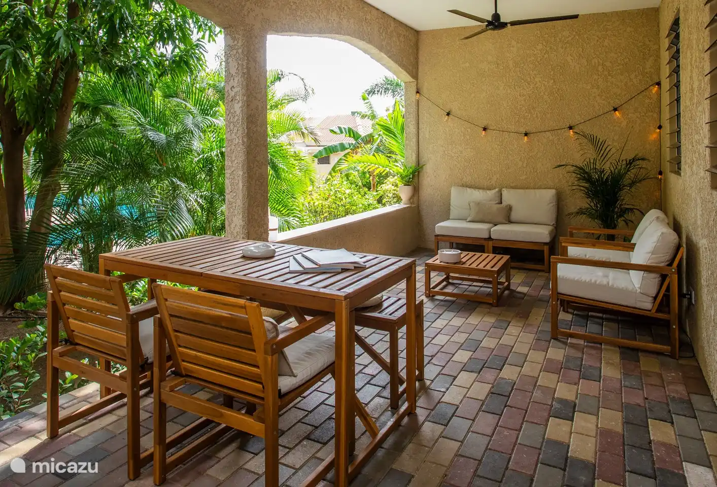 Porche spacieux au rez-de-chaussée avec accès direct à la piscine, abrité par des plantes tropicales et des arbres. Comprend un coin salon confortable, une table à manger et un ventilateur de plafond pour plus de confort