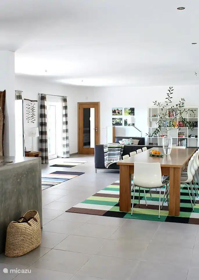 Das große Wohnzimmer mit durchgehenden Terrassentüren, die zur Veranda führen. Eine gemütliche Sitzecke am Holzofen und ein großer Esstisch mit Sitzgelegenheit für die ganze Familie.