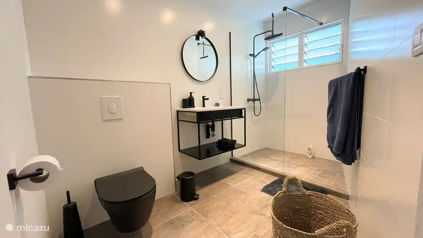 La salle de bain principale est accessible par la chambre principale et offre un double lavabo avec miroir, une douche à effet pluie et une douchette, des toilettes, une fenêtre à volets en aluminium pour une bonne ventilation, un sèche-cheveux et un panier à linge.