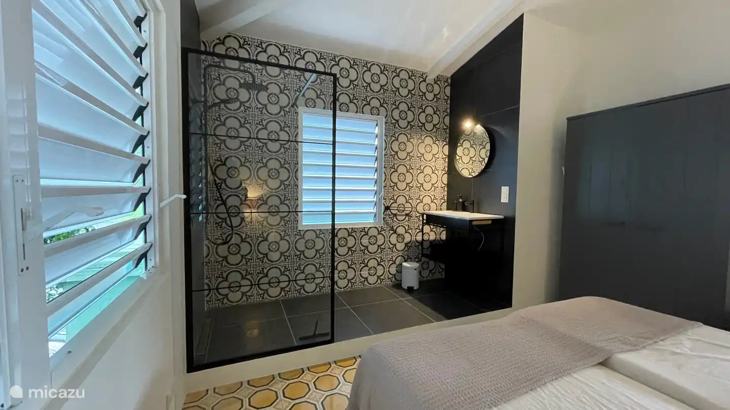 La salle de bain ouverte de la chambre 2 dispose d'une douche à effet pluie, d'une douchette, d'un lavabo avec miroir, d'un panier à linge et de la climatisation.