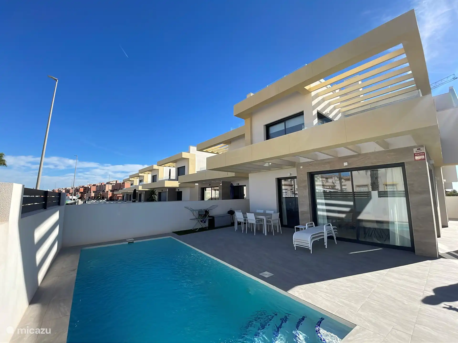 Blick auf die Villa, einschließlich Poolbereich und Terrasse mit Liegestühlen und Esstisch im Freien. Der Bereich ist sowohl von der Küche als auch vom Wohnzimmer aus zugänglich.