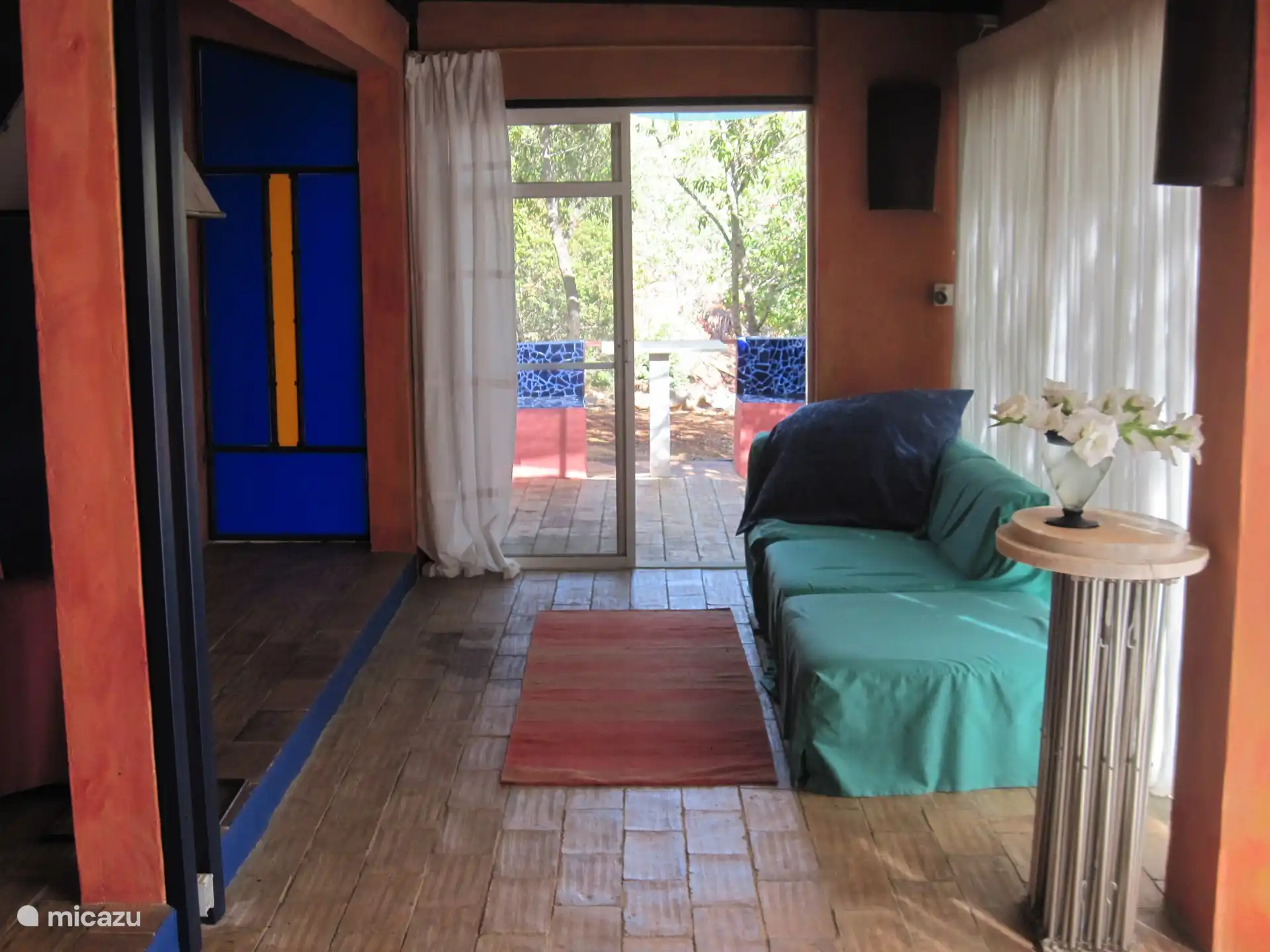 Blick in den farbenfrohen Lebensraum mit Sofa und angrenzender Schlafzimmertuer und einem  hinteren Ausgang auf eine kleine Terrasse mit Sitzbank.