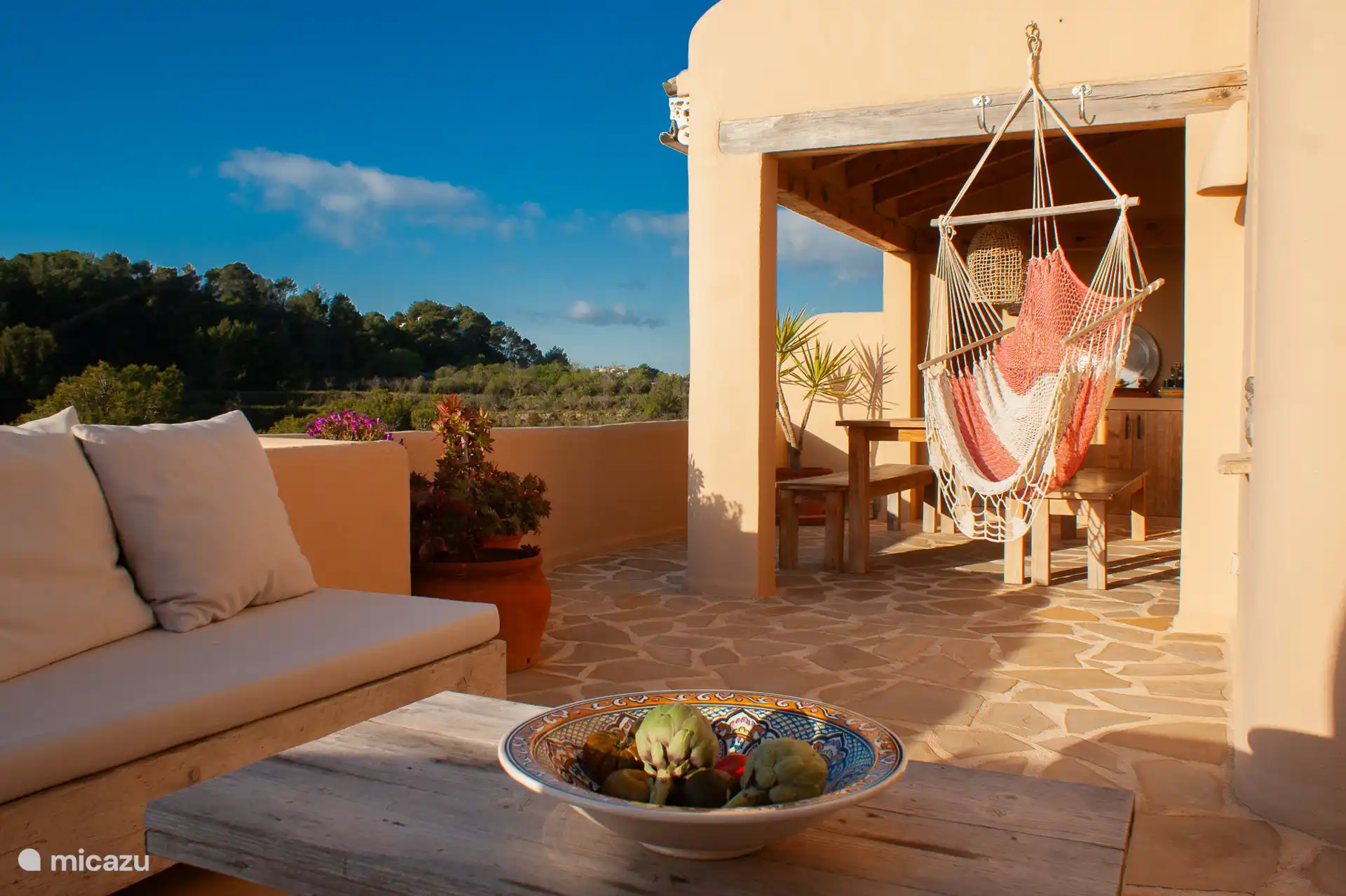 Overdekt terras op de begane grond met prachtig uitzicht, een ruime eettafel voor 6 personen, een comfortabele zithoek met loungekussens, schommelstoel en een buitenkeuken.
