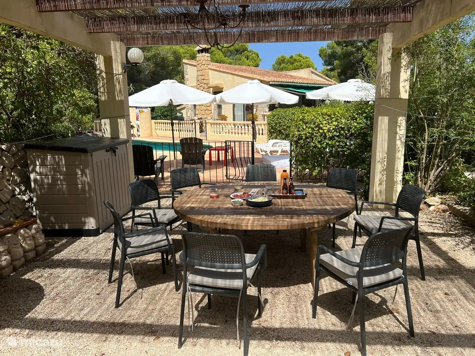 Ronde tafel onder de pergola in de semi schaduw met uitzicht op het zwembad en daarachter het huis.