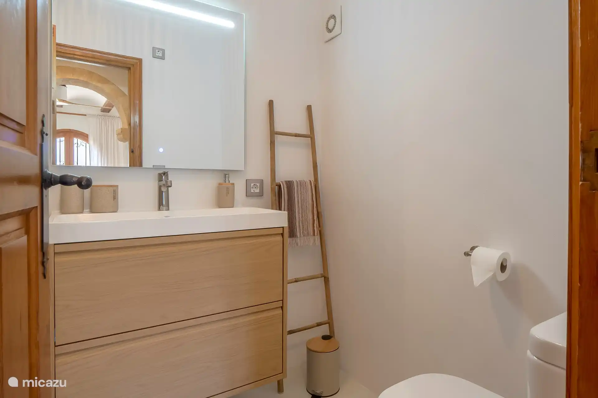 Das Badezimmer mit ebenerdiger Dusche, WC, Dunstabzugshaube, Waschbecken und Spiegel mit LED-Beleuchtung