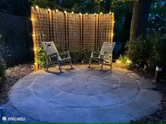 Ein Sitzbereich mit natürlichen Pflanzen und geflochtenen Weidenmatten bietet sowohl Privatsphäre als auch eine warme Atmosphäre. Die Ambientebeleuchtung rund um das Gitter sorgt für ein intimes Ambiente, während die Schaukelstühle und der Tisch zu entspannten Abenden bei einem Glas Wein einladen.