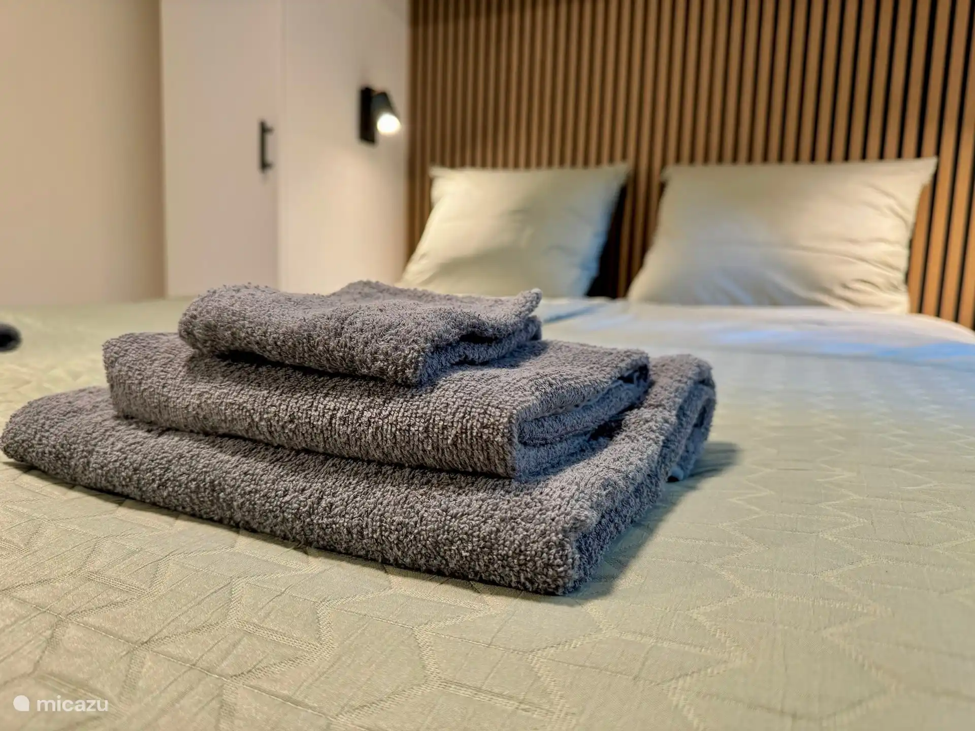 Das Hauptschlafzimmer versprüht in diesem Wellness-Ferienhaus modernen Luxus mit einem bequemen Boxspringbett, einer warmen Holzlattenwand mit schwarzem Hintergrund und stilvollen Leselampen auf beiden Seiten. während USB-A- und USB-C-Anschlüsse am Bett zusätzlichen Komfort bieten.