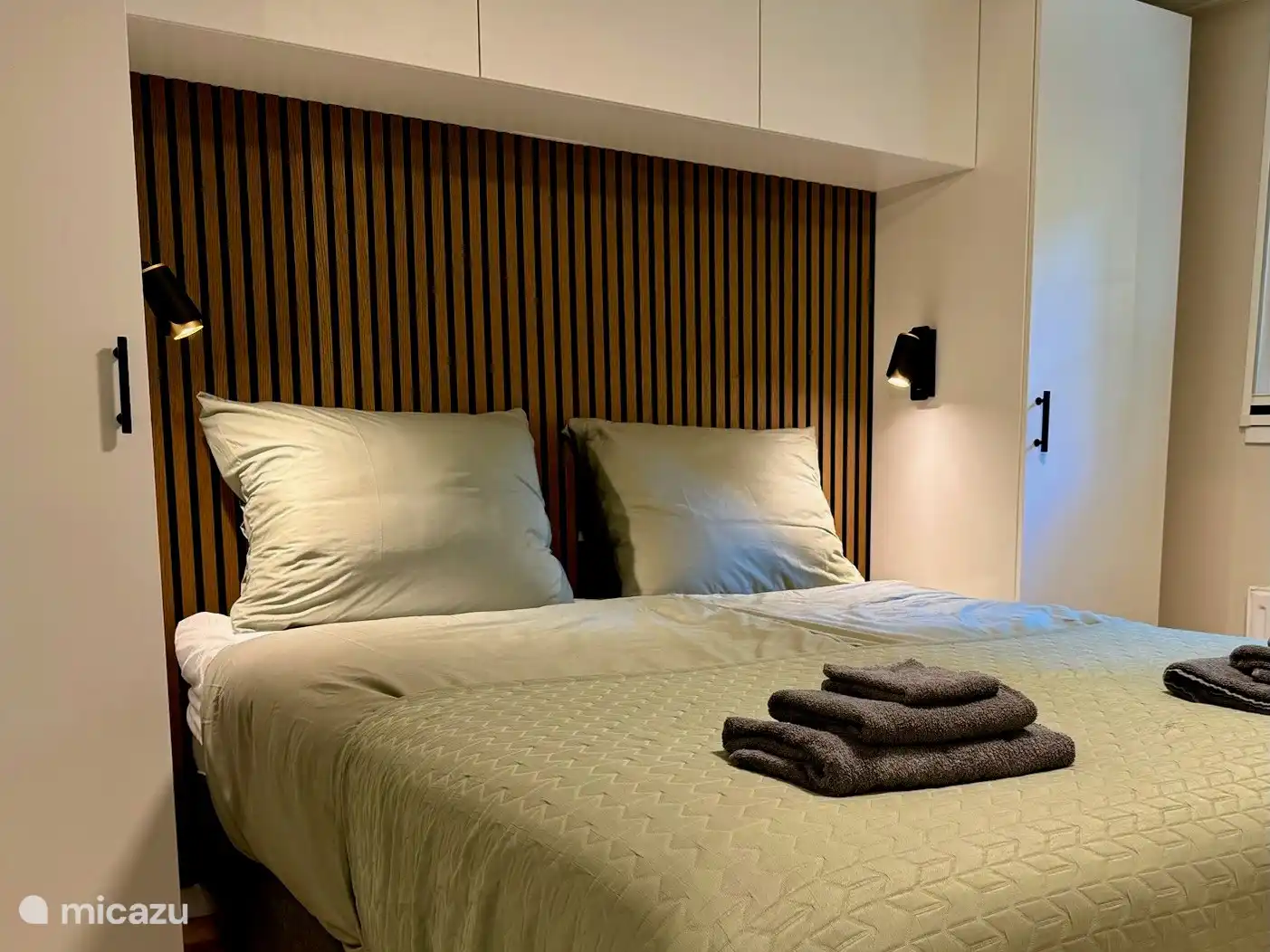 Das Hauptschlafzimmer ist elegant nach zeitgenössischen Standards gestaltet. Akzentuiert wird das komfortable Boxspringbett durch eine warme Holzlattenwand mit schwarzem Hintergrund und akustischem PET-Filz. Die weißen Umrüstschränke links und rechts bieten geräumigen, beleuchteten Stauraum. 