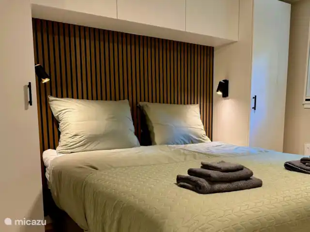 Maison de vacances Maridu Family Wellness | Pays-Bas, Gueldre, Hattemerbroek - chalet La chambre principale est élégamment conçue selon les normes contemporaines. Le confortable lit à sommier tapissier est accentué par une paroi à lattes de bois chaleureuse sur fond noir et un feutre PET acoustique. Les armoires de conversion blanches à gauche et à droite offrent un espace de rangement spacieux et éclairé.