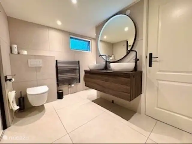 Maison de vacances Maridu Family Wellness | Pays-Bas, Gueldre, Hattemerbroek - chalet Le mobilier de salle de bains allie style et fonctionnalité avec une finition gris-brun naturel. Équipé de deux lavabos blancs, de robinets noirs modernes et d’un grand miroir rond avec éclairage annulaire. Des prises de courant sous le miroir et un sèche-cheveux dans le tiroir complètent ce meuble.