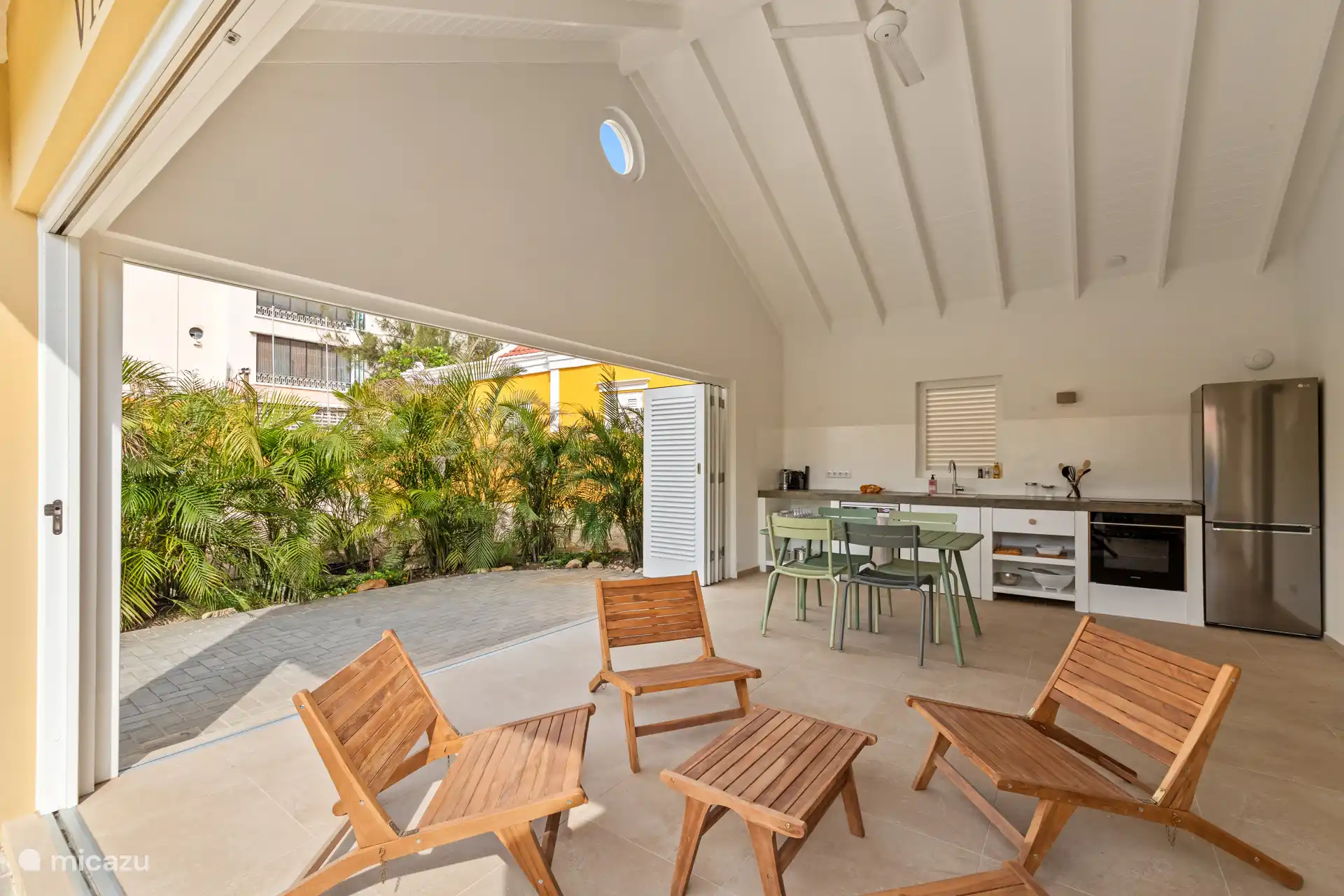 Villa Blenchi with open kitchen, dining table and sitting area under high ceiling with ceiling fan. Large sliding doors connect the interior with the tropical greenery and the spacious terrace