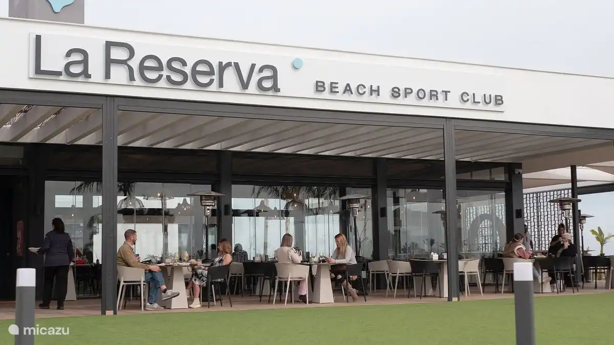 Une photo du Beach Resort Club montre un espace de restauration animé et accueillant au bord de la plage. L’espace est meublé avec des tables en bois élégantes et des chaises confortables, parfaites pour les clients qui souhaitent profiter d’un déjeuner ou d’un dîner tranquille.