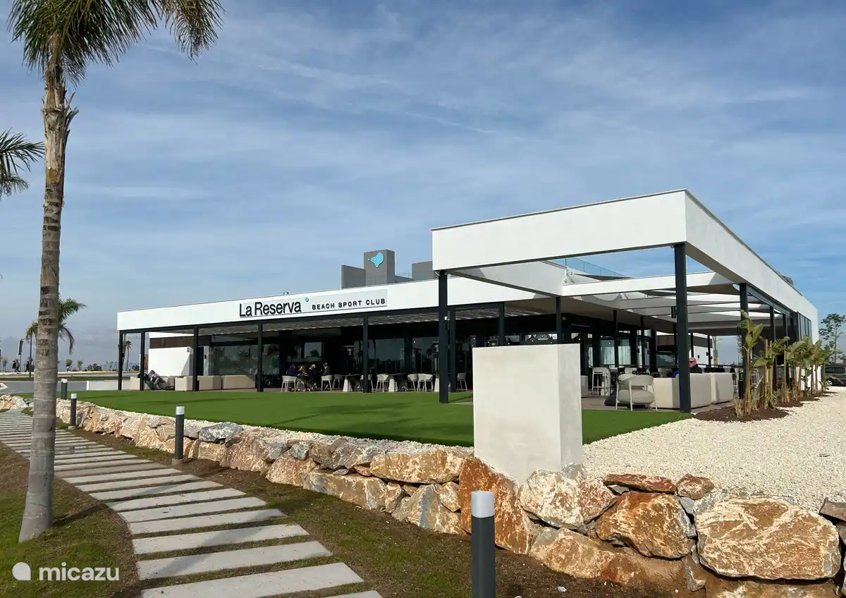 Une photo du Beach Resort Club montre un espace de restauration animé et accueillant au bord de la plage. L’espace est meublé avec des tables en bois élégantes et des chaises confortables, parfaites pour les clients qui souhaitent profiter d’un déjeuner ou d’un dîner tranquille.