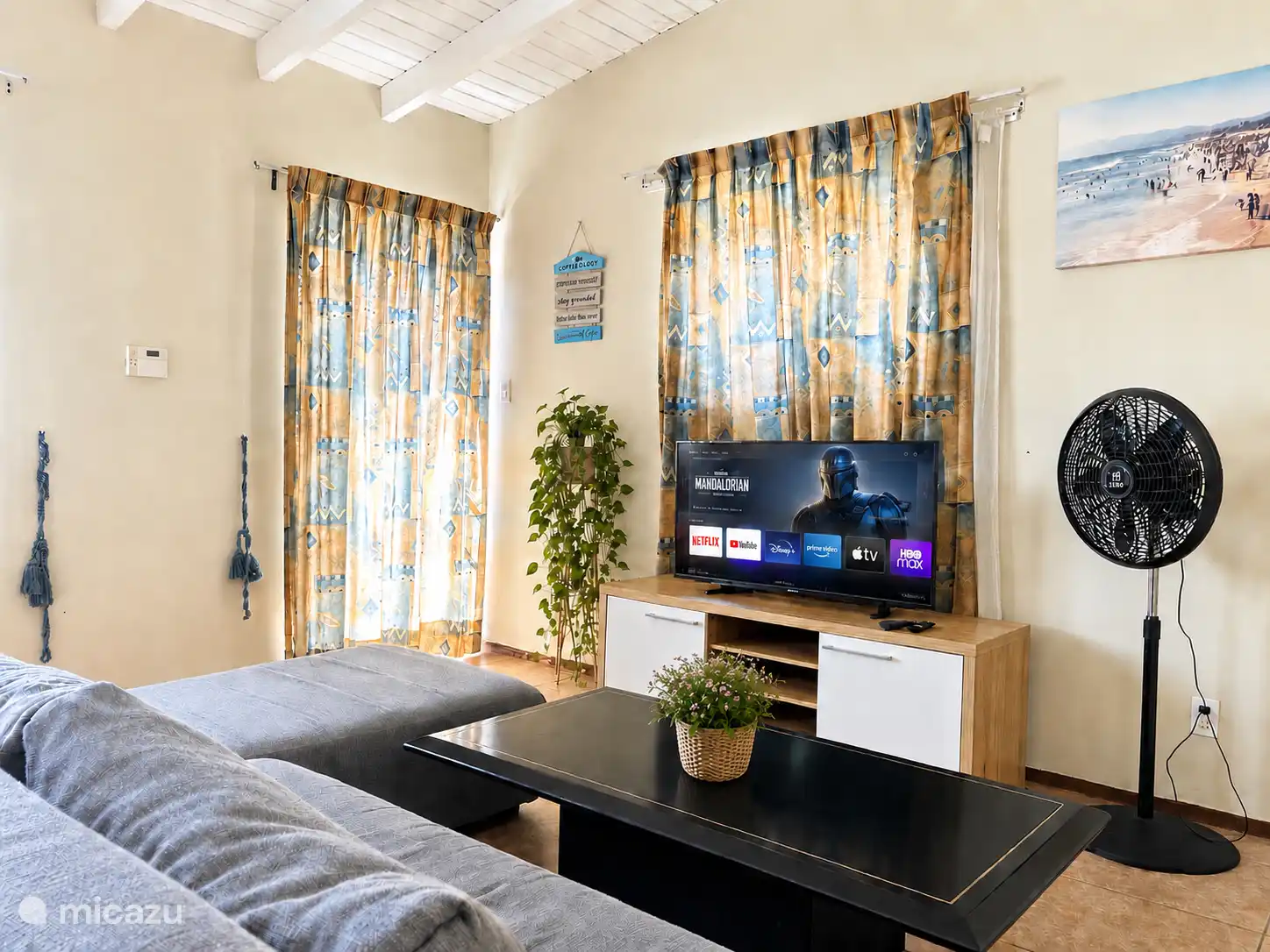Ruime en comfortabele woonkamer met een flatscreen-tv, ventilator en een gezellige L-vormige bank. De kamer heeft meerdere ramen en deuren die zorgen voor veel frisse lucht.