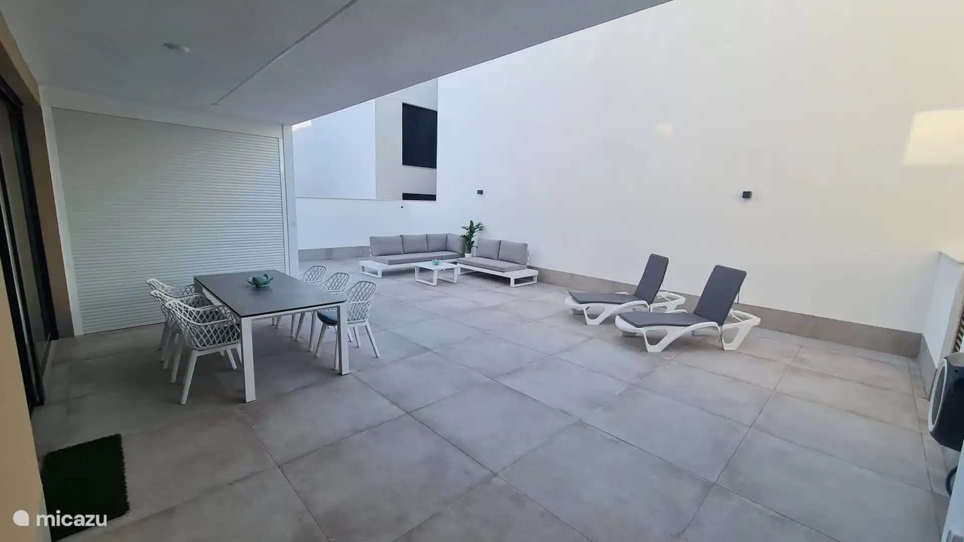 De la partie couverte de la terrasse, une vue sur l’ensemble de la terrasse. Sous le baldaquin, une table pour six personnes avec des chaises et des coussins. Sur la terrasse un coin salon. 