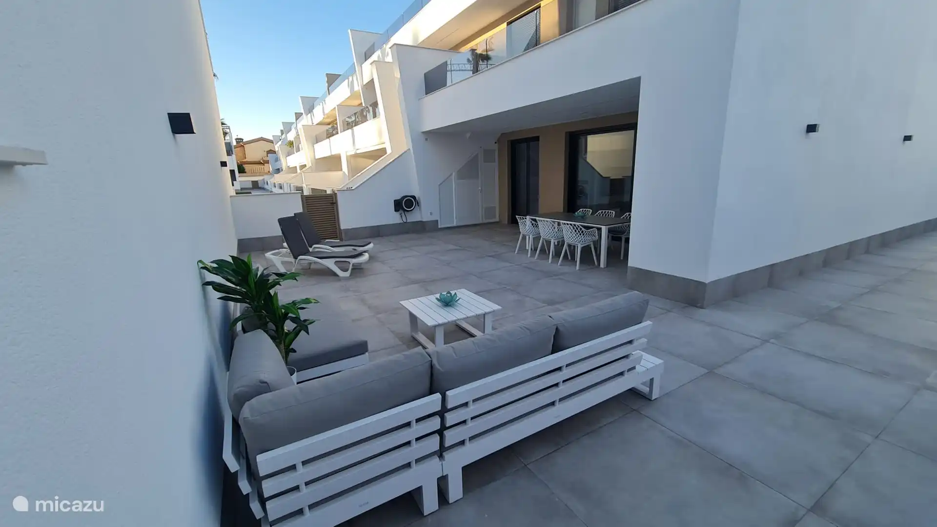 Photo d’ensemble de la terrasse avec salon et table à manger. Porte qui donne accès à la piscine commune. Machine à laver disponible dans le débarras à l’extérieur. 