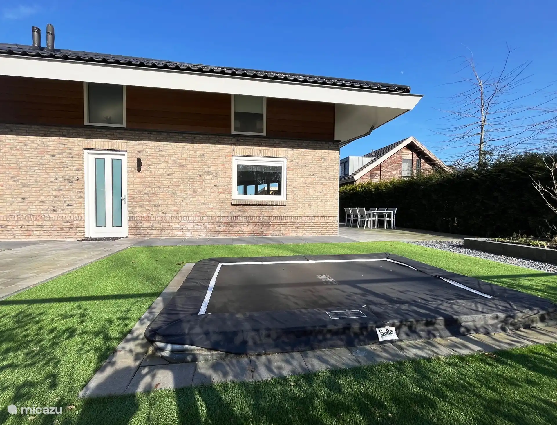 Vor dem Haus, mit einer hohen Hecke und einem Tor, das vom Hof getrennt ist. Die Kinder können sich auf dem Trampolin austoben.