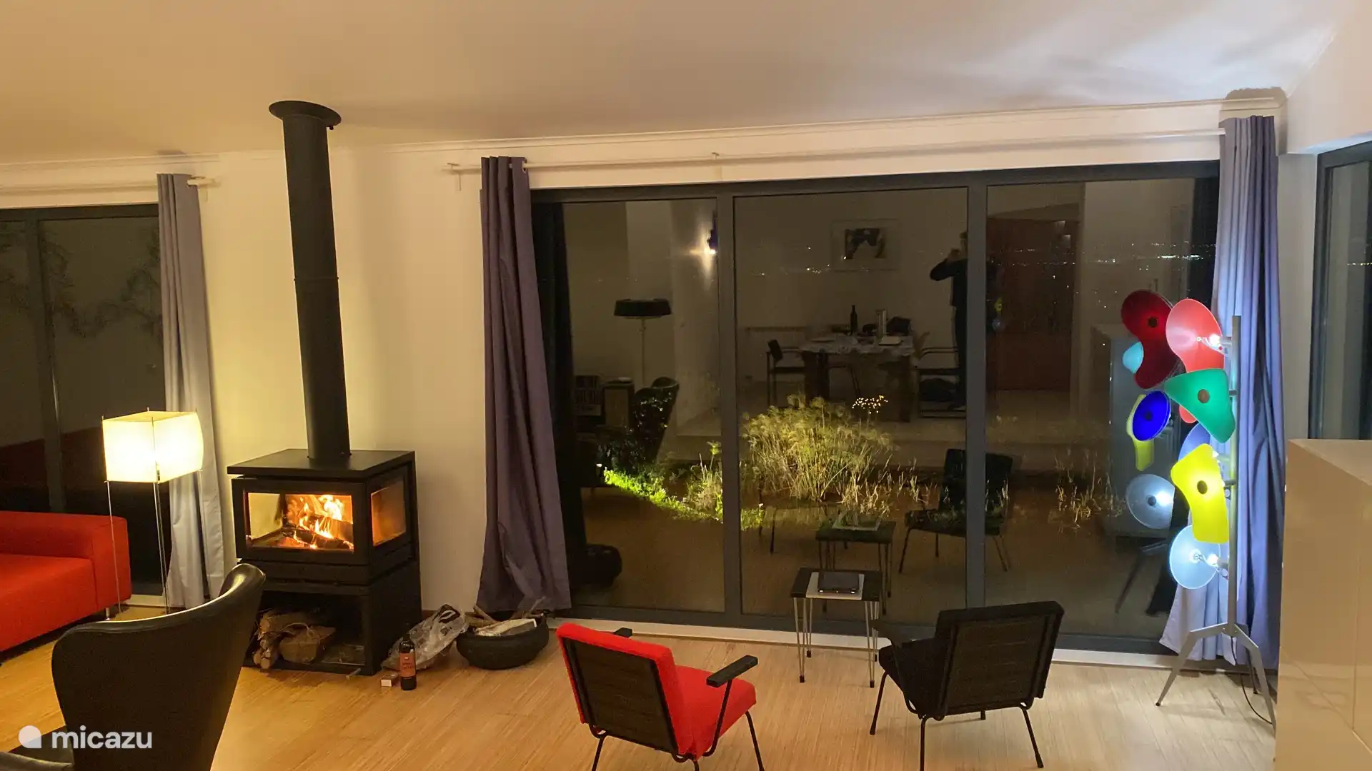 Huiskamer ‘s-avonds met brandende houtkachel en verlichte vijver