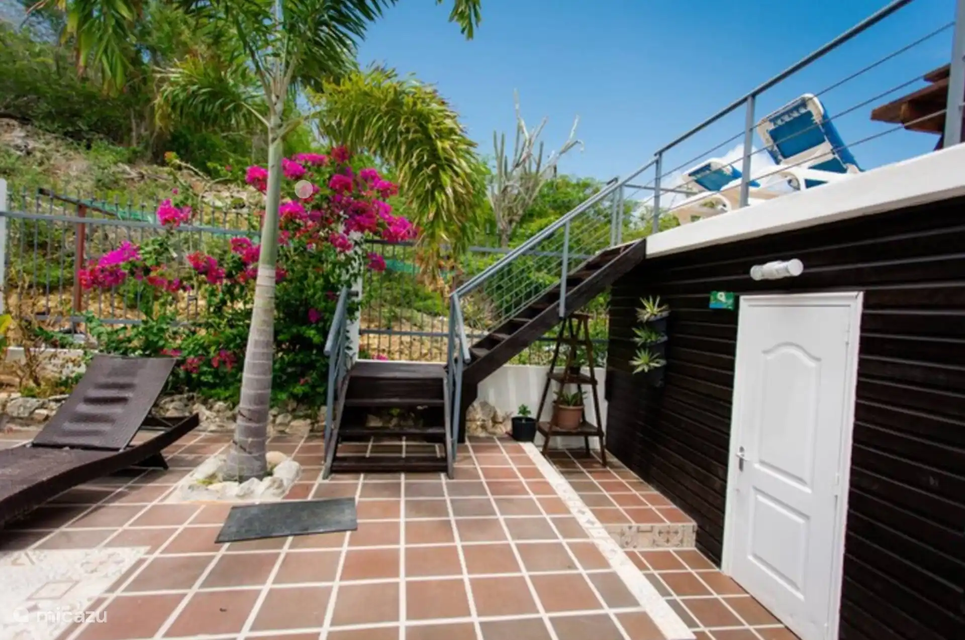 Über eine charmante Treppe gelangt man auf die Sonnenterrasse auf dem Dach oberhalb des Studiokomplexes. Palmen, blühende Bougainvillea und ein Lounge-Bett schaffen eine entspannte, natürliche Atmosphäre.