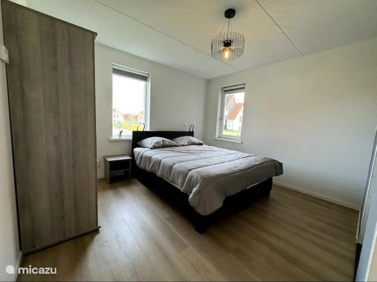 Lichte slaapkamer waar daglicht door twee ramen naar binnen stroomt. De warme houtvloer en zachte grijstinten creëren een rustige, moderne plek om volledig tot ontspanning te komen.