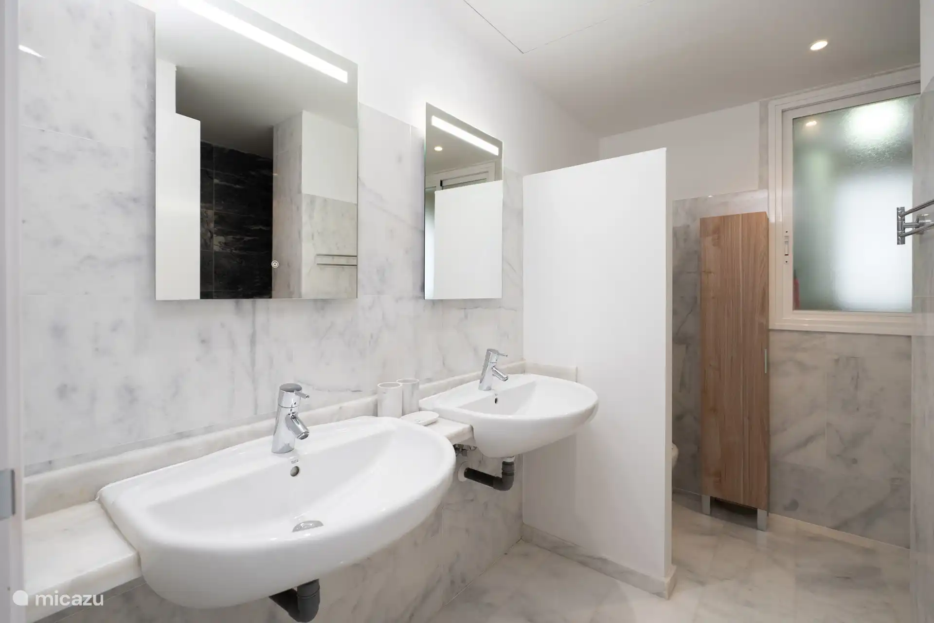 Salle de bain avec double vasque, miroirs éclairés, toilettes, bidet et un placard de rangement haut.
