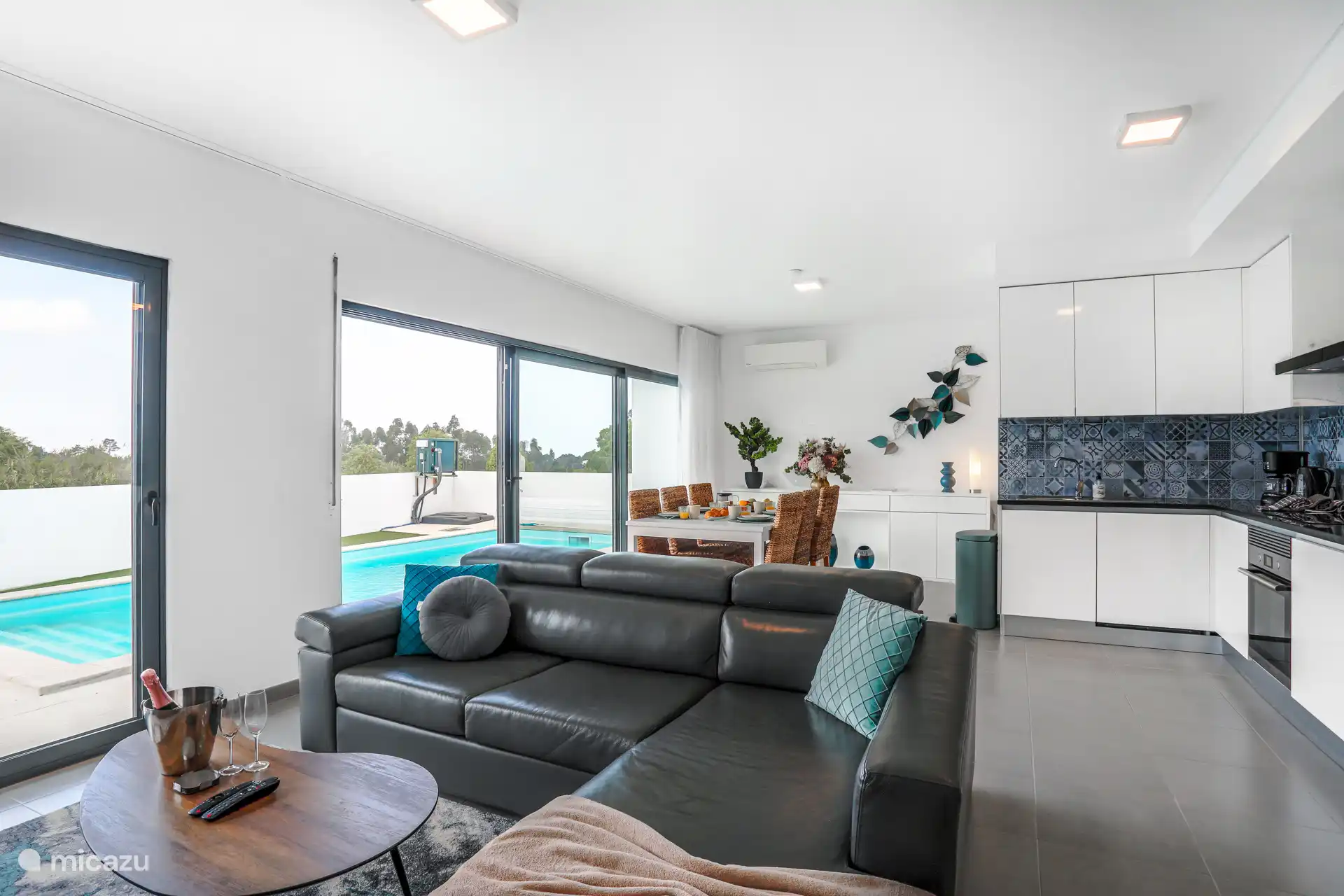 Vue d’ensemble du salon avec une grande quantité de lumière et une vue sur la piscine. Toute la façade - ainsi que toutes les fenêtres de la villa - est équipée de volets. 