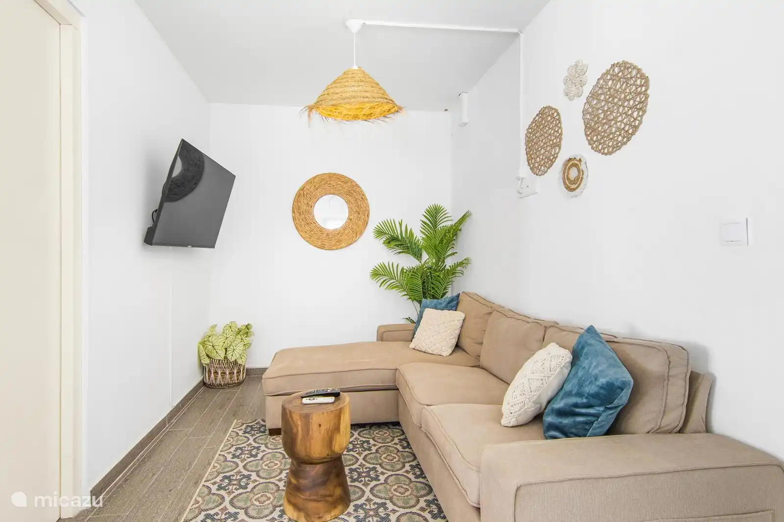 Acogedora sala de estar con sofá beige, cojines tejidos y azules, planta tropical y espejo de mimbre, creando un ambiente fresco y relajante.