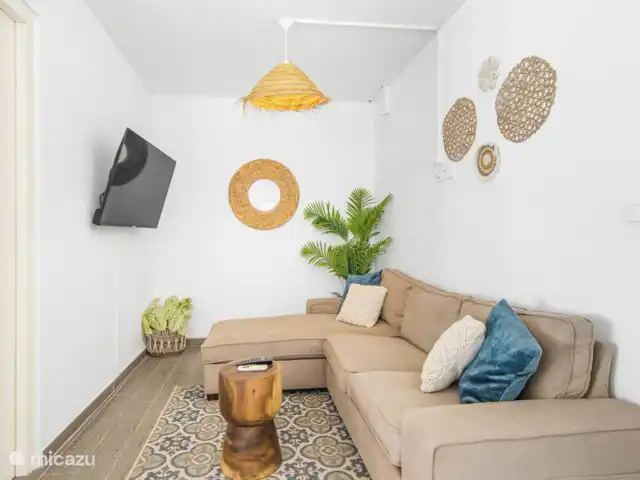 SOMETHING ELS huren in Curaçao, Banda Ariba (oost), Marie Pampoen - vakantiehuis Acogedora sala de estar con sofá beige, cojines tejidos y azules, planta tropical y espejo de mimbre, creando un ambiente fresco y relajante.