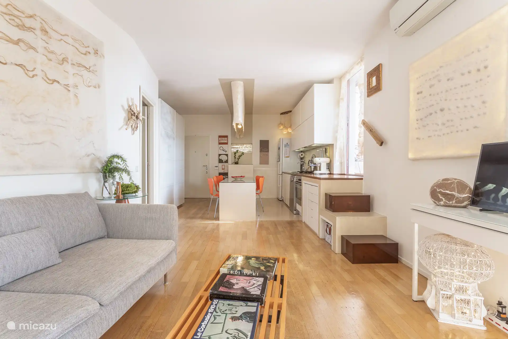 Amplia sala de estar y cocina de planta abierta con aire acondicionado, Smart TV y acceso al balcón y la terraza a través de cuatro escalones.