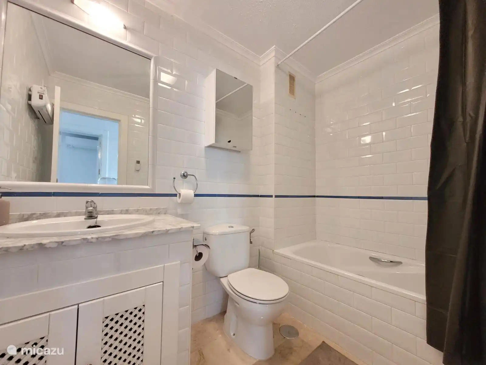 Salle de bain avec baignoire, douche, WC et lavabo avec rangements. Les carreaux blancs et la ligne d’accent bleue donnent à la pièce un aspect frais et soigné.